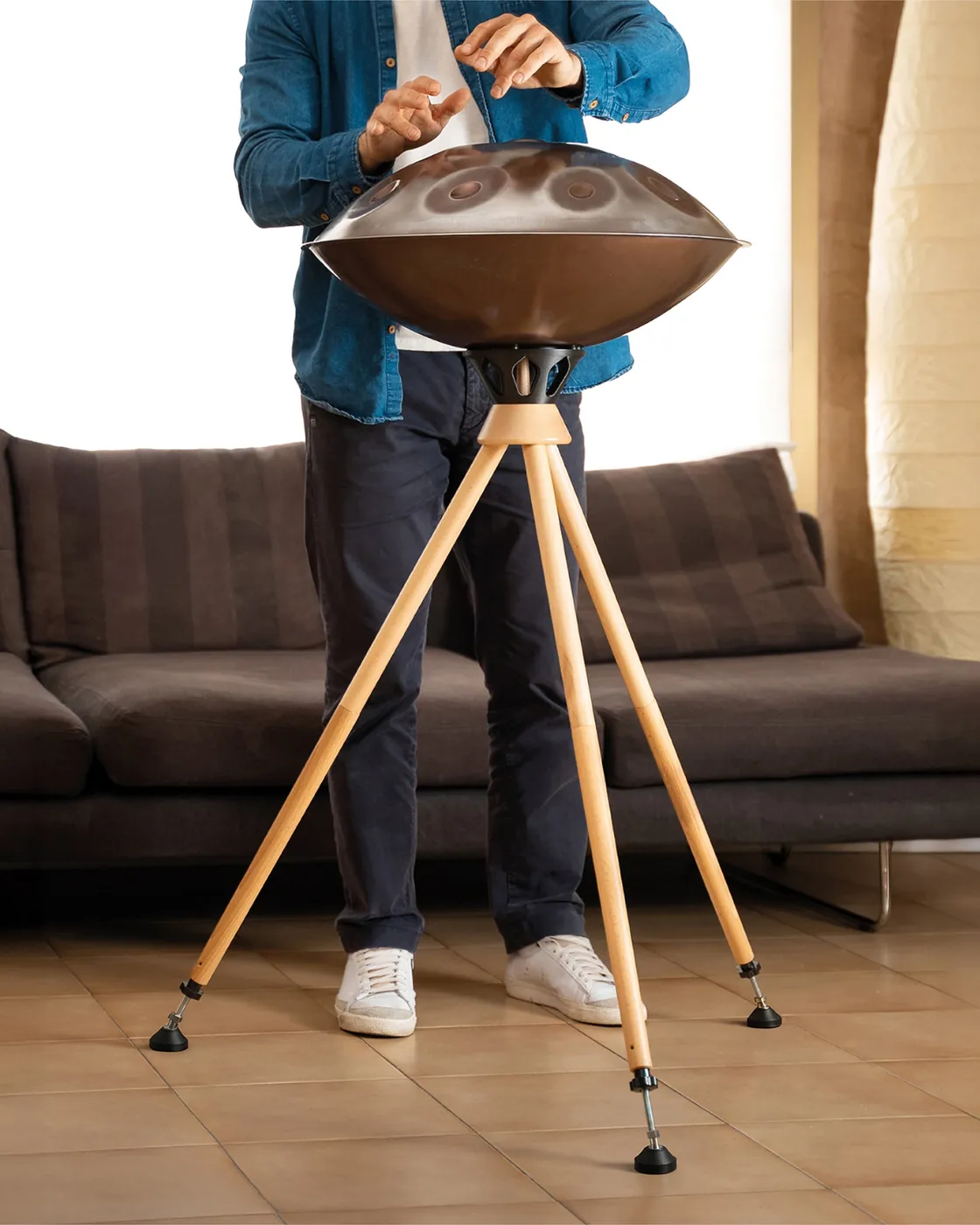 Un musicien joue debout sur un handpan posé sur le trépied Atlas All, dans un salon : les trois pieds en bois clair sont déployés en position haute.